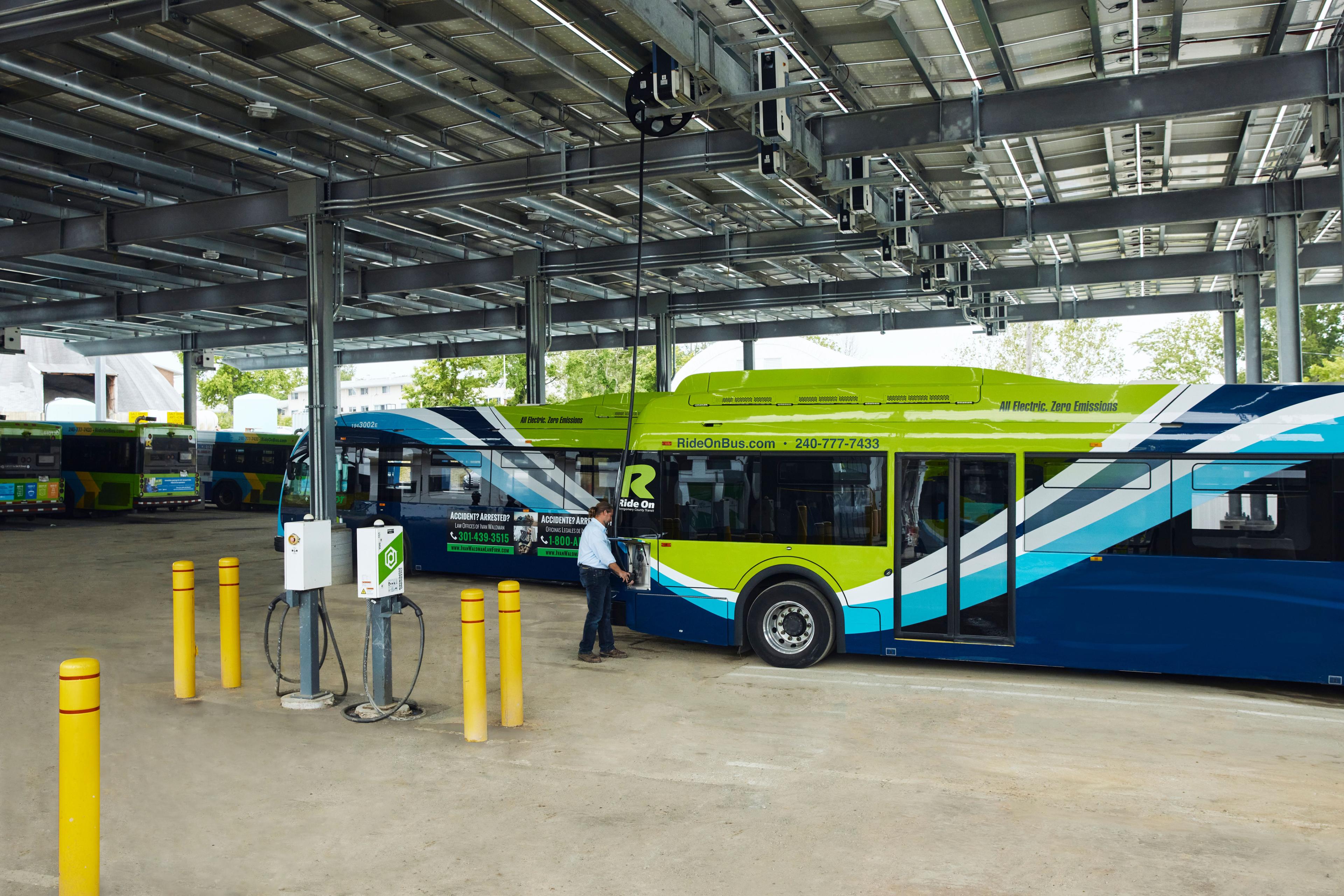 A man plugs in an electric bus under a canopy of solar panels.