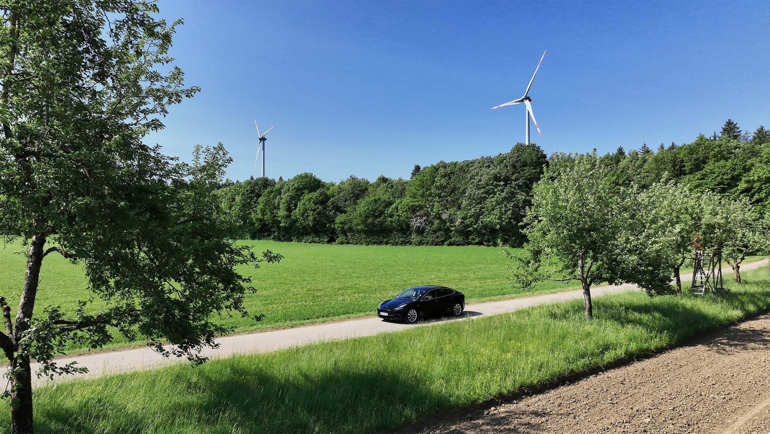 Tesla fährt auf einer Landstraße mit Windrädern im Hintergrund