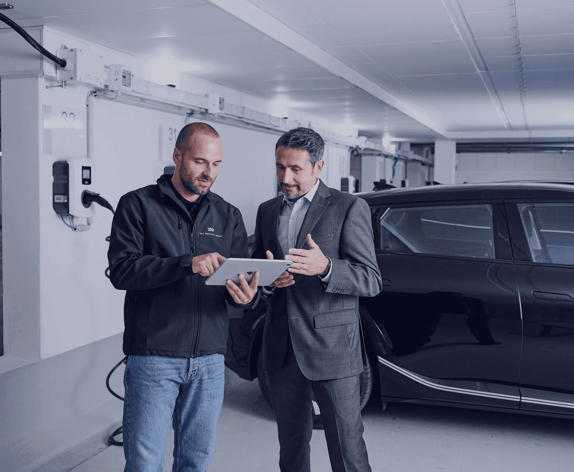 Elektriker und Kunde schauen gemeinsam auf Tablet in der Hand mit ladendem Elektroauto im Hintergrund