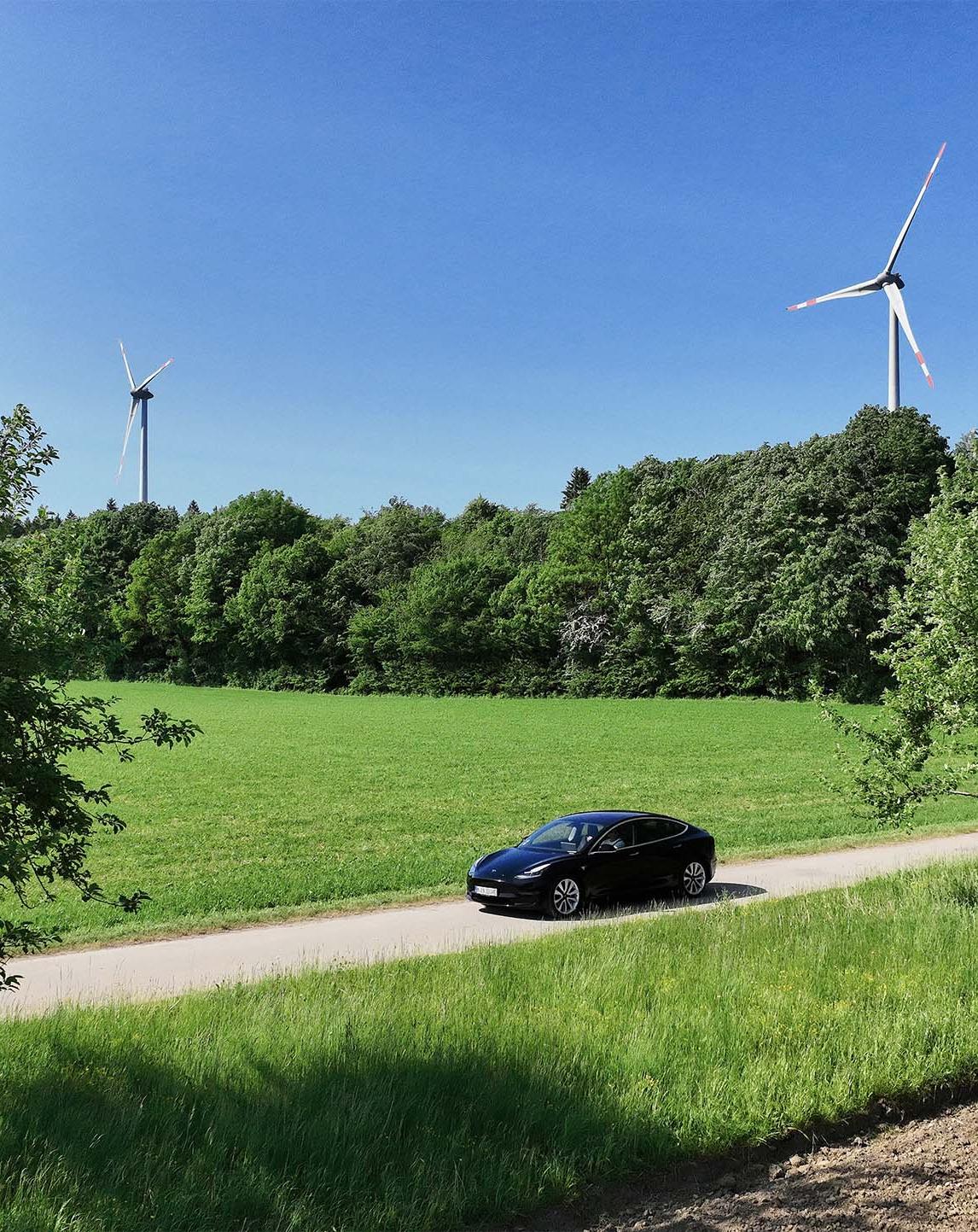 Tesla fährt auf einer Landstraße mit Windrädern im Hintergrund