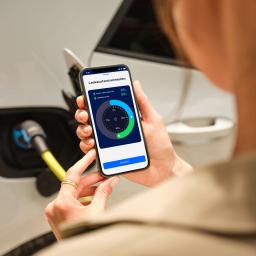 Eine Frau blickt auf ihr Handy vor einem ladenden Elektroauto