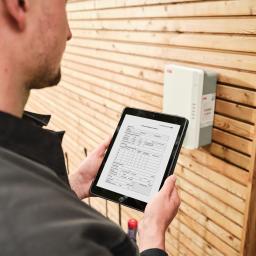 Ein Mann steht vor einer Wallbox an einem Holzhaus und haelt ein Tablet in den Haenden